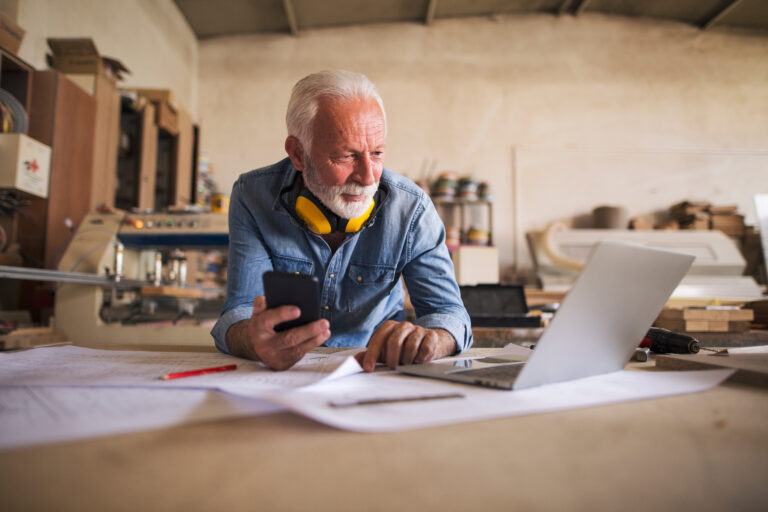 Seitenansicht eines Handwerkers, der am Schreibtisch sitzt, ein Mobiltelefon hält und auf den Laptop schaut